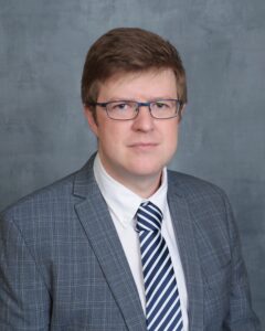 Headshot of Daniel Cooper wearing glasses, a grey suit and blue and white striped tie.