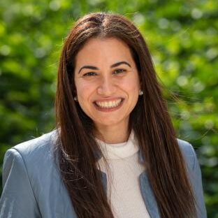 Headshot with long, brown hair, smiling and a white top and blue jacket.