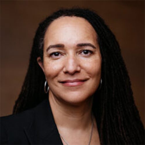 Headshot, smiling wearing a dark suit with long hair and earrings.