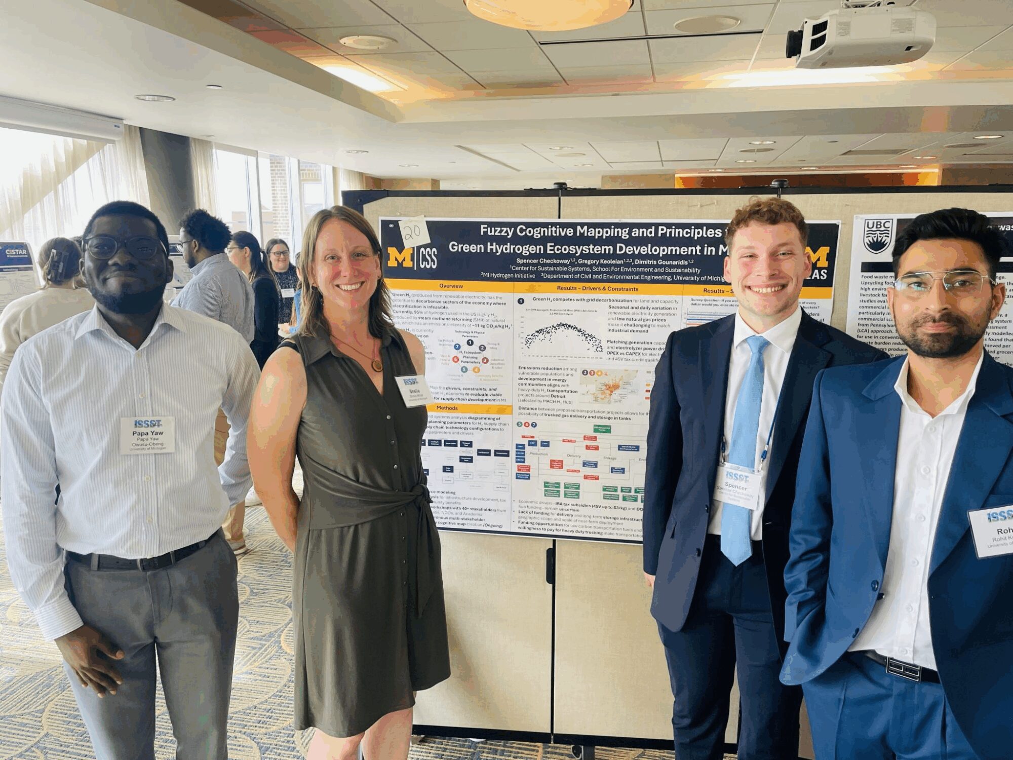 Four people are standing in front of a scientific poster presentation at a conference. One individual, wearing a light-colored shirt, has a name tag that reads