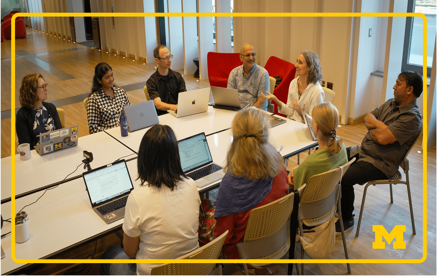 Group of people sit in a classroom around tables with laptops, engaged in active discussion and collaborative work