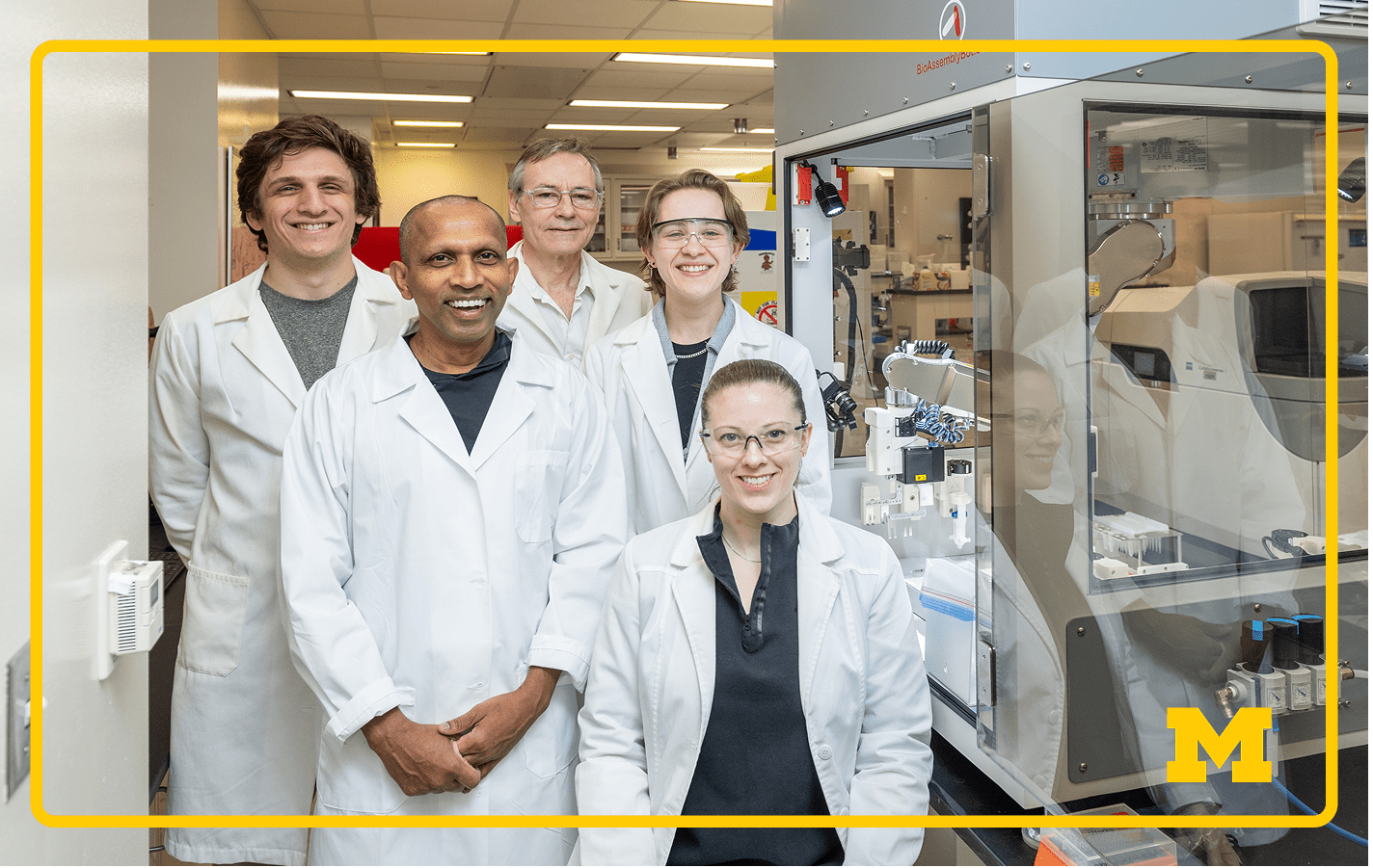 Front Left to Right: Indika Rajapakse, Lindsey Muir, “BAB” Back Left to Right: Joshua Pickard, Walter Meixner, Jillian Cwycyshyn lab coats pose inside a modern laboratory, standing next to BioAssemblyBot 400 machine.