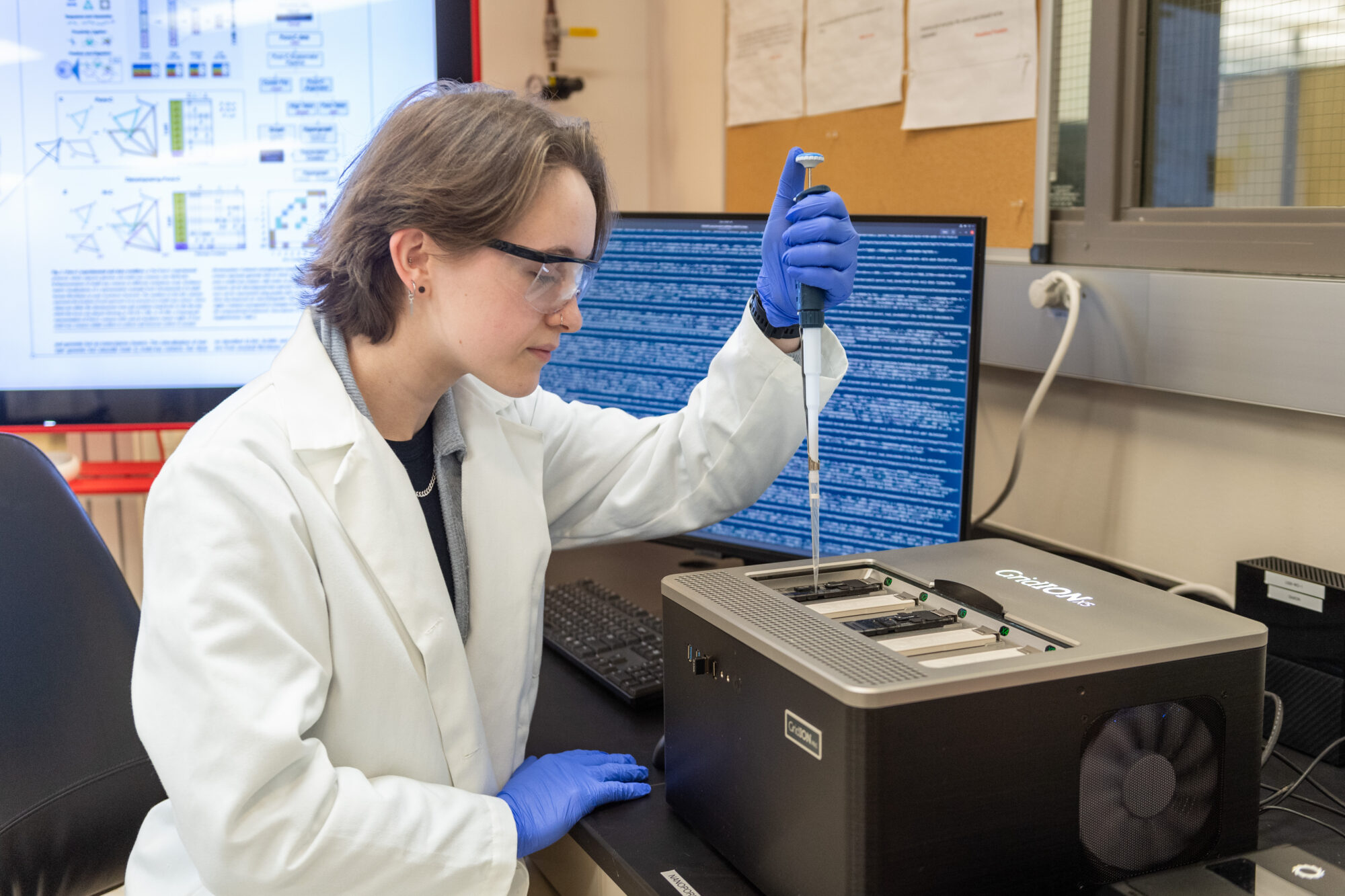 Jillian Cwycyshyn preparing cells for sequencing Jillian Cwycyshyn in a lab coat and gloves prepares cells for sequencing with advanced equipment and research displays nearby