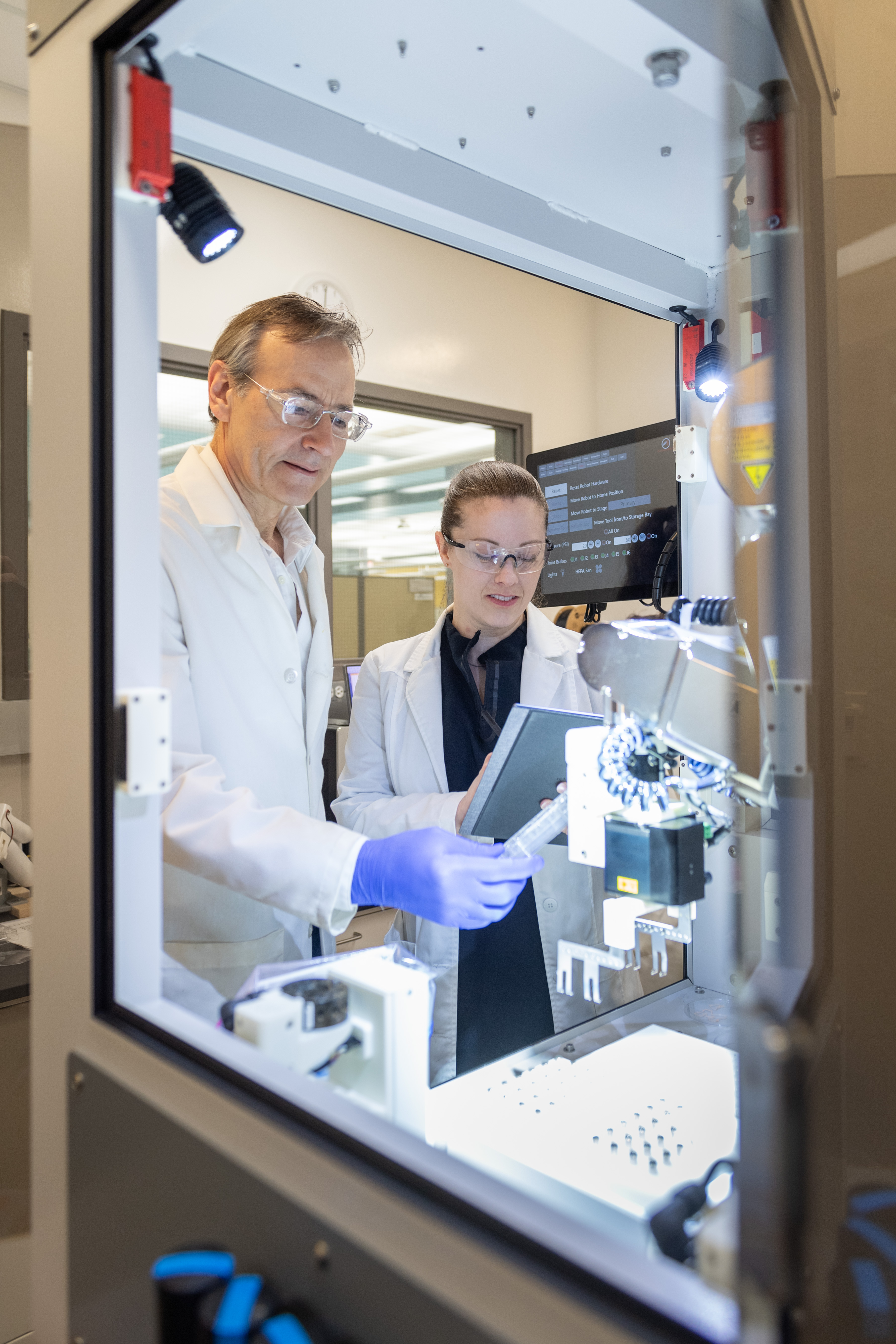 Lindsey Muir and Walter Meixner using BAB Lindsey Muir and Walter Meixner in lab coats operate the BioAssemblyBot 400 machine, examining samples and recording data on a tablet.