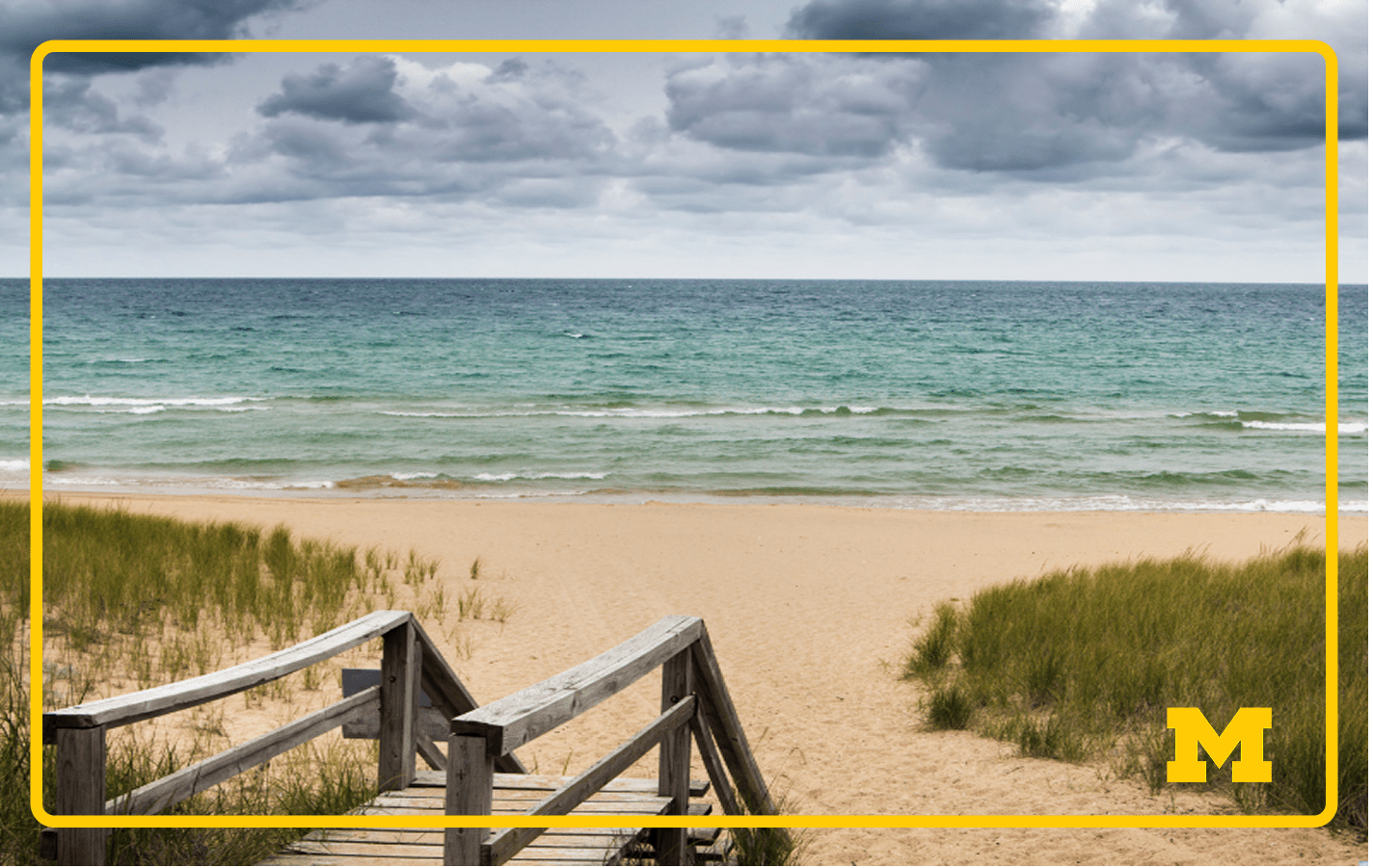 Protect great lakes science Wooden stairs leading to a sandy beach with grassy dunes, under a cloudy sky and calm ocean waves.