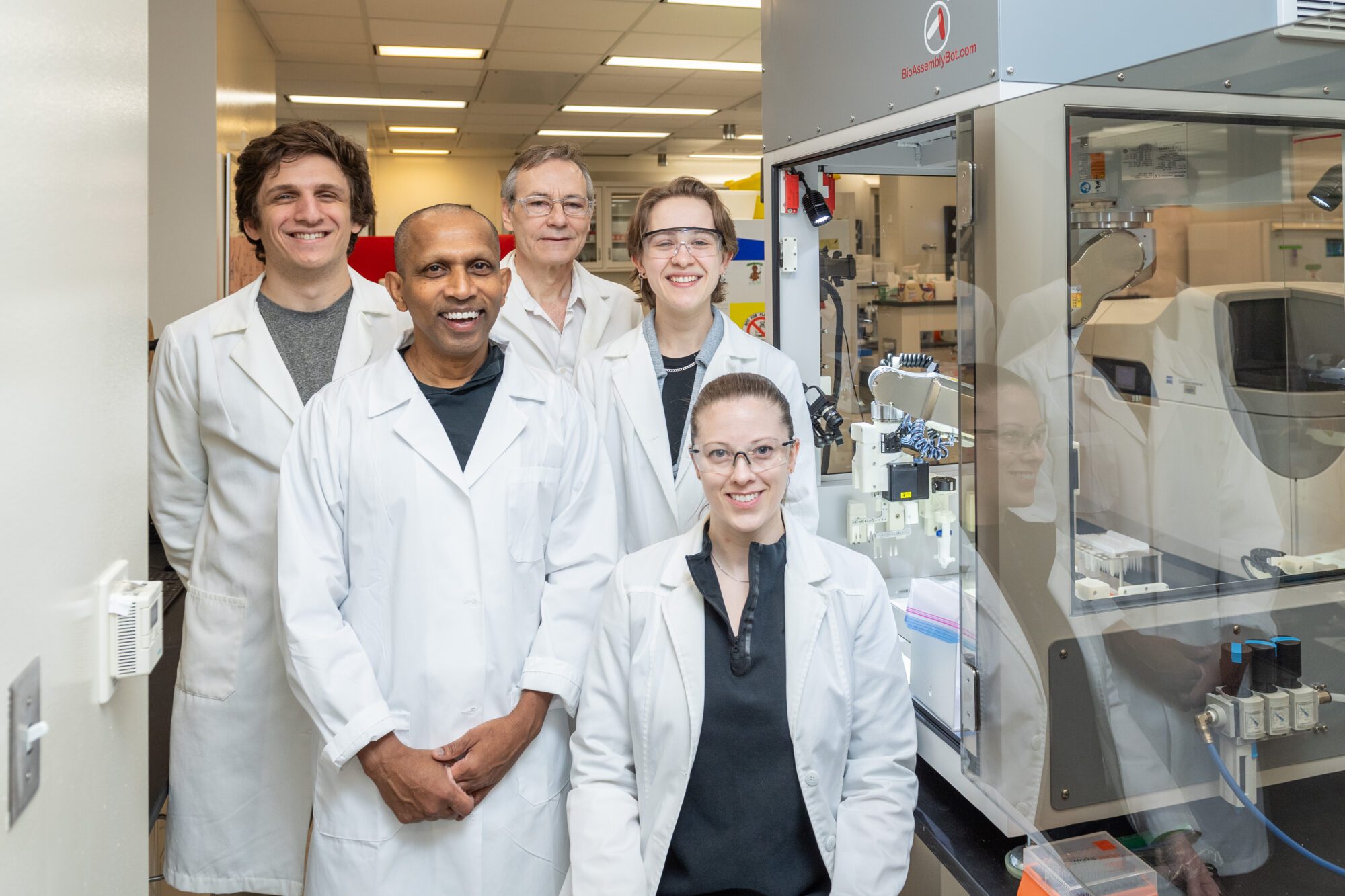 A group photo of people in a research lab, wearing lab coats. Front Left to Right: Indika Rajapakse, Lindsey Muir, Back Left to Right: Joshua Pickard, Walter Meixner, Jillian Cwycyshyn