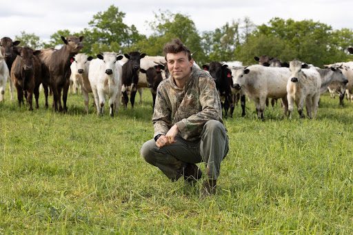 Will Brinkerhoff, a PhD candidate at the University of Michigan School for Environment and Sustainability (SEAS) Will Brinkerhoff in camouflage clothing squats on green grass in a pasture, with a herd of cattle standing behind him.