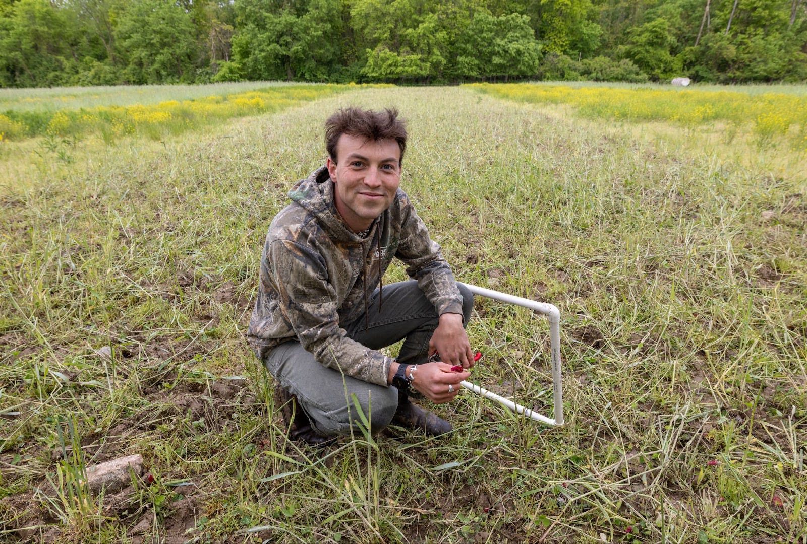 Will Brinkerhoff, a PhD candidate at the University of Michigan School for Environment and Sustainability (SEAS) Will Brinkerhoff in camouflage jacket kneeling in a grassy field, holding scissors and a white PVC square sampling frame.