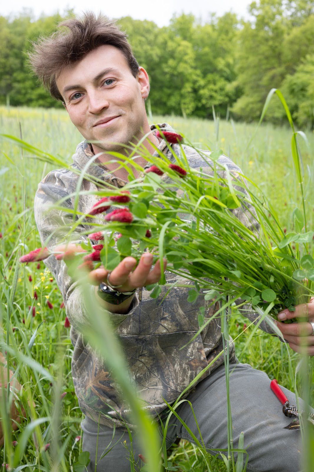 Will Brinkerhoff, a PhD candidate at the University of Michigan School for Environment and Sustainability (SEAS) Will Brinkerhoff in camouflage clothing presenting a handful of leafy stems with small red flowers in a grassy field.