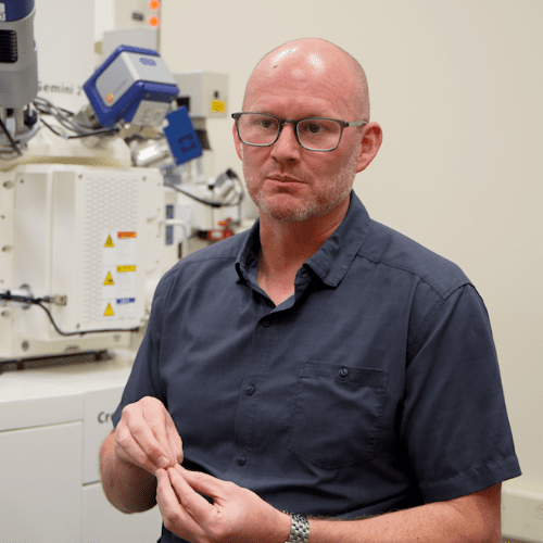 A researcher in a dark shirt stands in front of advanced microscopy equipment, holding an object and explaining laboratory details.