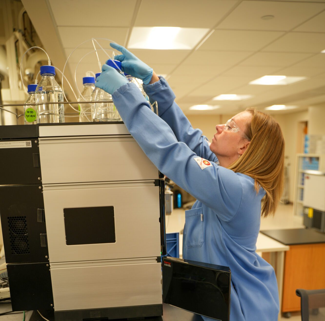 A lab technician in a blue coat adjusts bottles connected to a scientific instrument in the lab.