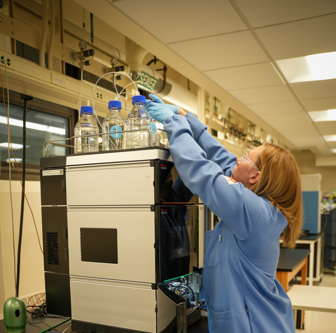 A technician wearing a blue lab coat sets up a scientific instrument with connected bottles and tubes.
