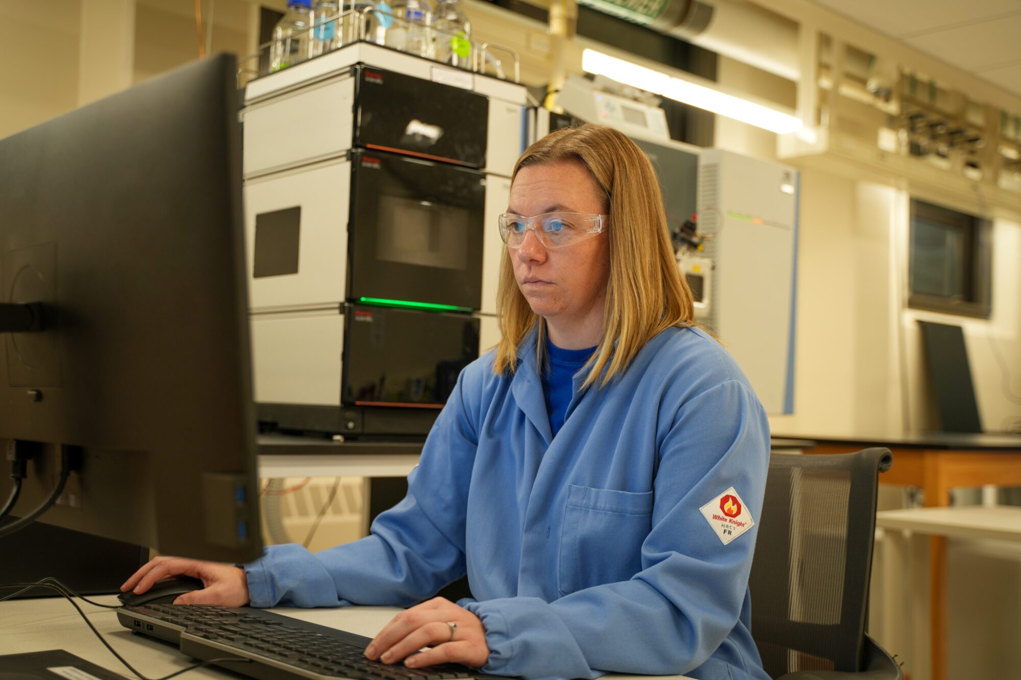 A person wearing a blue lab coat works at a computer in a laboratory with scientific equipment in the background.