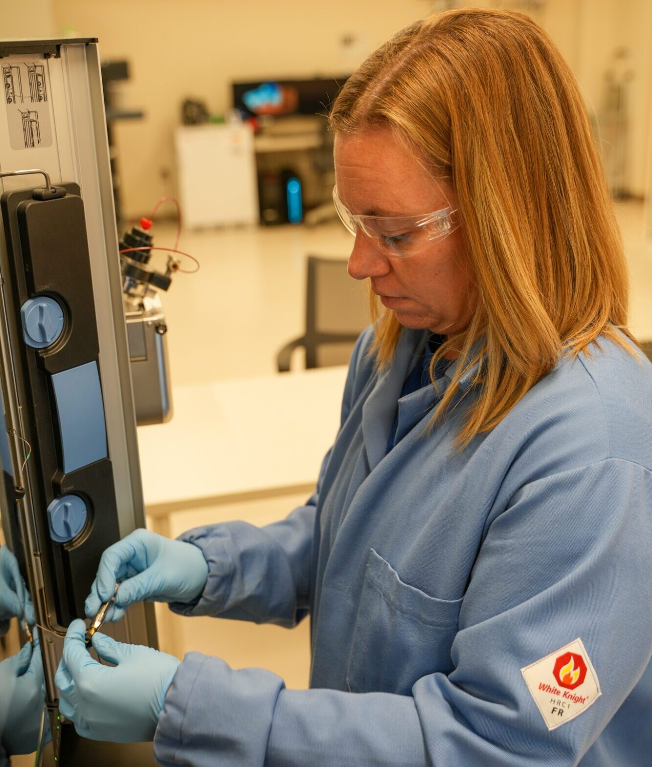 A person in a blue lab coat and gloves uses scientific instrumentation in a laboratory setting with various equipment.