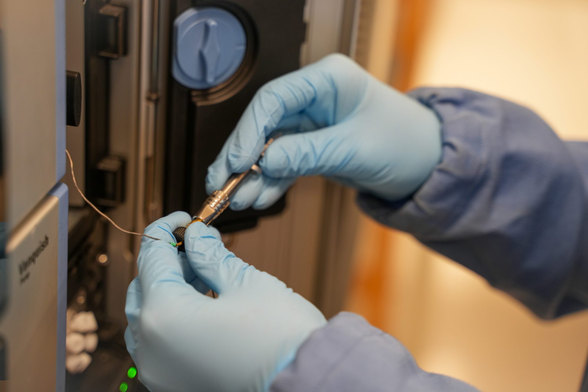 A person in a blue lab coat uses laboratory equipment, connecting wires with gloved hands in a scientific setting.