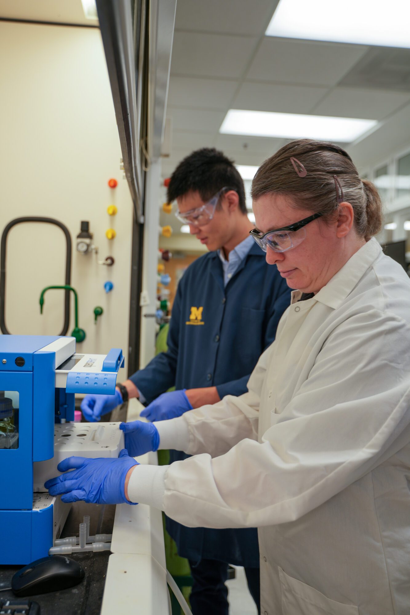 Two scientists in laboratory coats and gloves prepare and load samples into a device, surrounded by lab equipment.