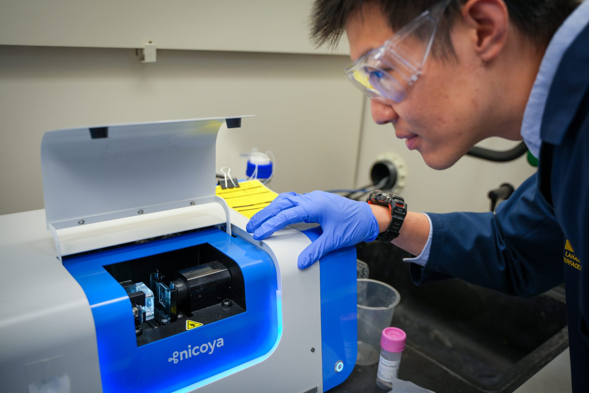 A scientist in blue gloves closely observes and interacts with a laboratory instrument on a benchtop setting.