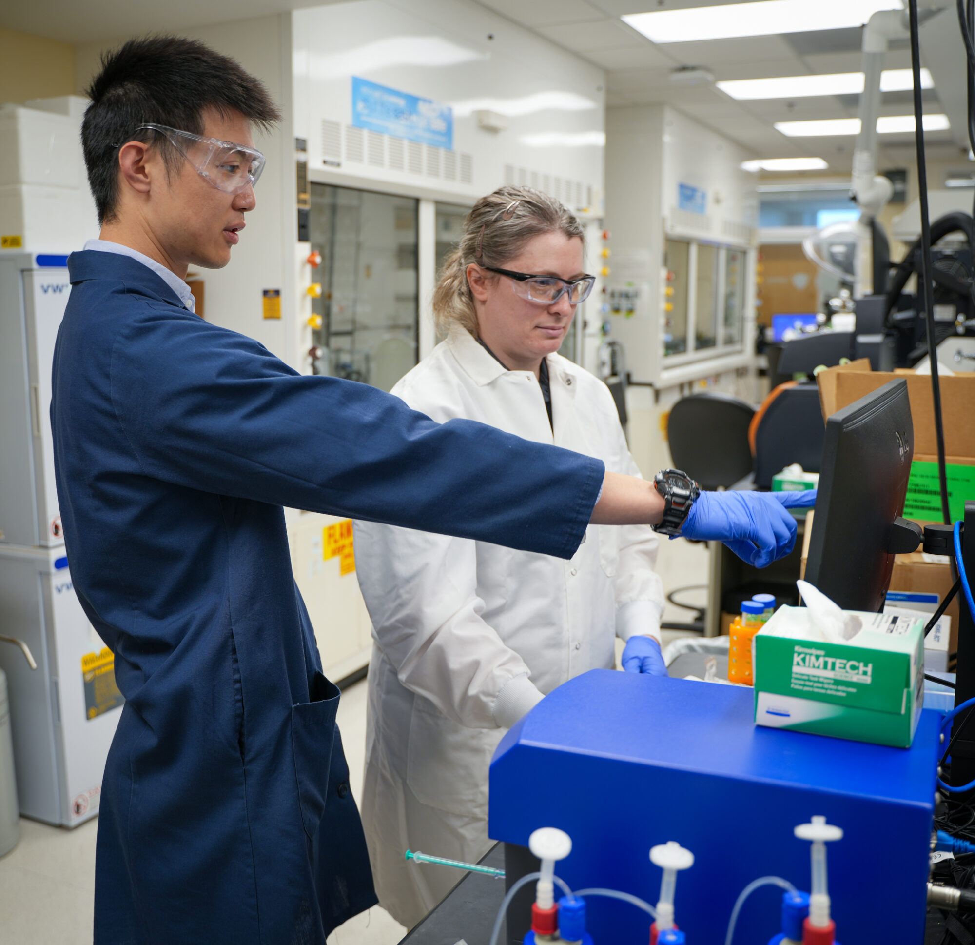 Two scientists wearing lab coats and safety goggles discuss data on a computer screen in a laboratory.