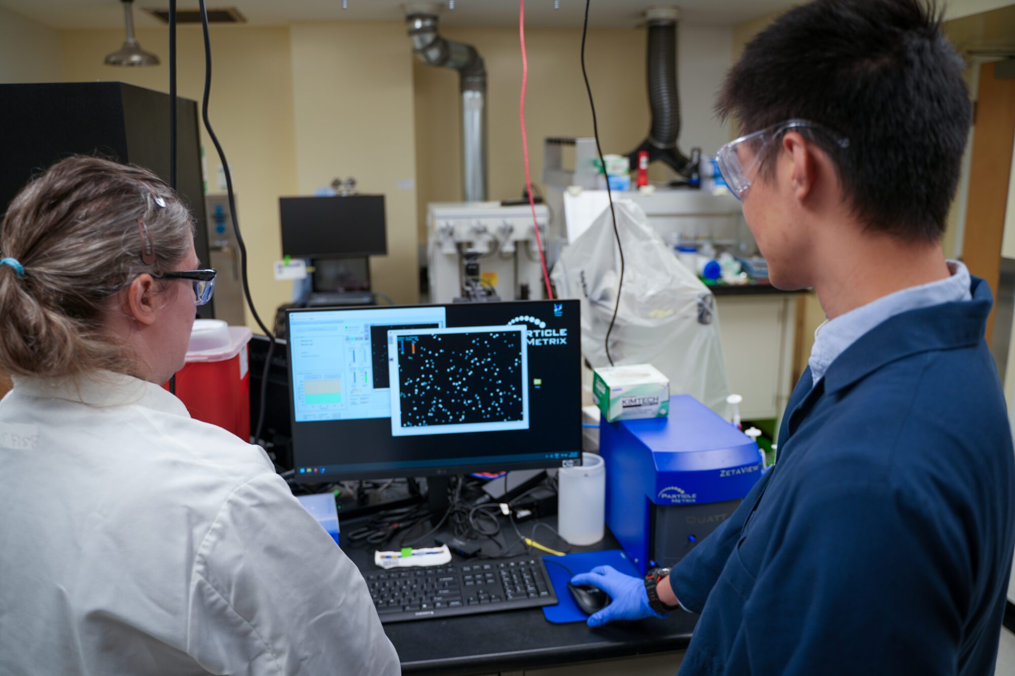 Researchers analyze particle tracking data displayed on a computer connected to lab equipment in a research lab.