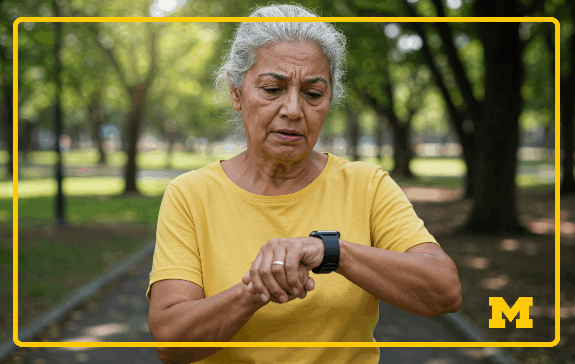 An older adult with gray hair wearing a yellow shirt checks their smartwatch while standing on a park path.