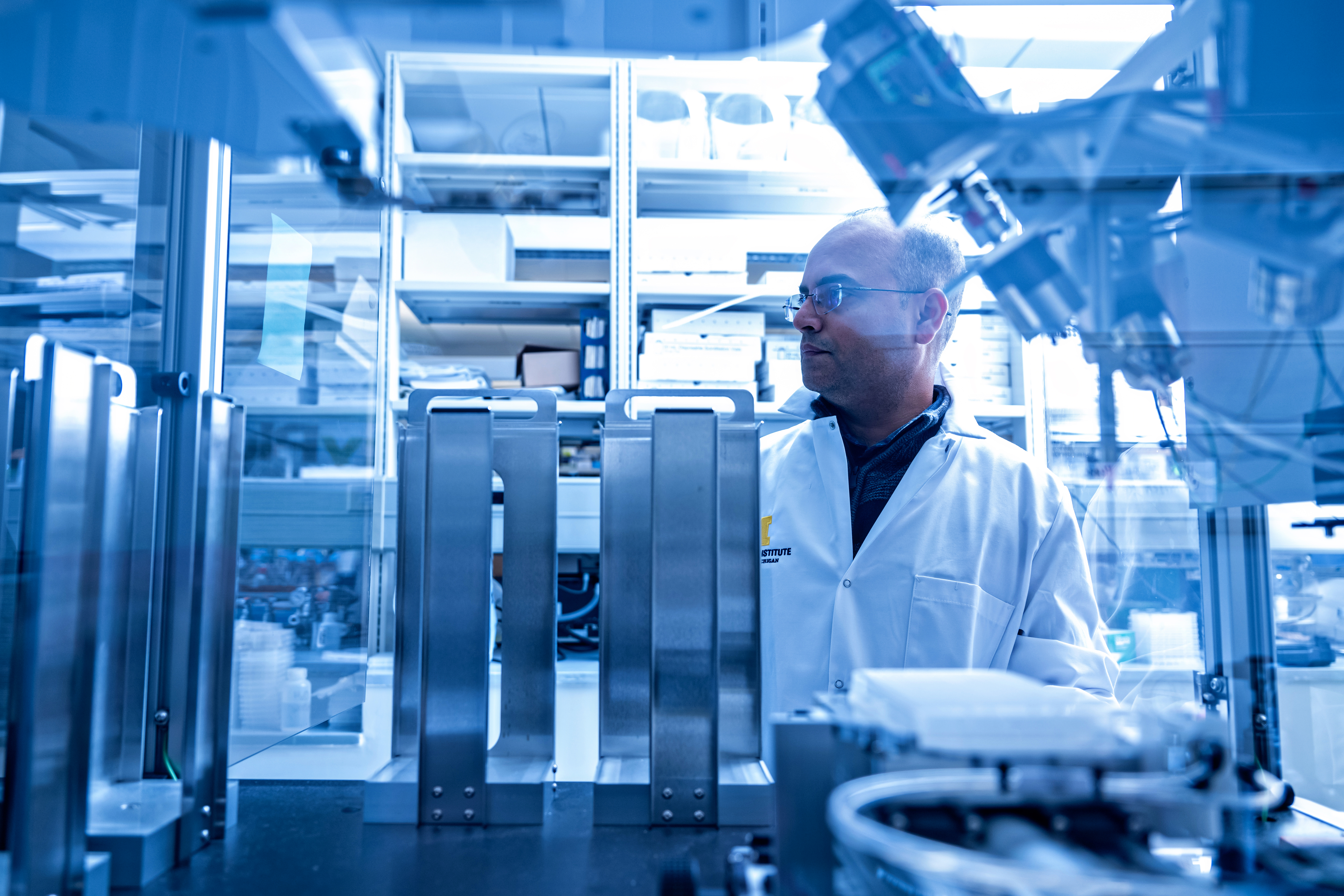 Scientist stands among advanced robotics and scientific instruments in a high-tech laboratory, shelves of equipment in background.
