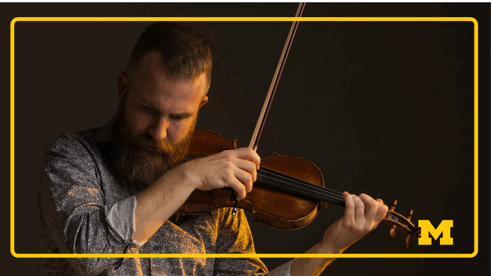 A bearded man playing the violin and looking off camera at the ground.