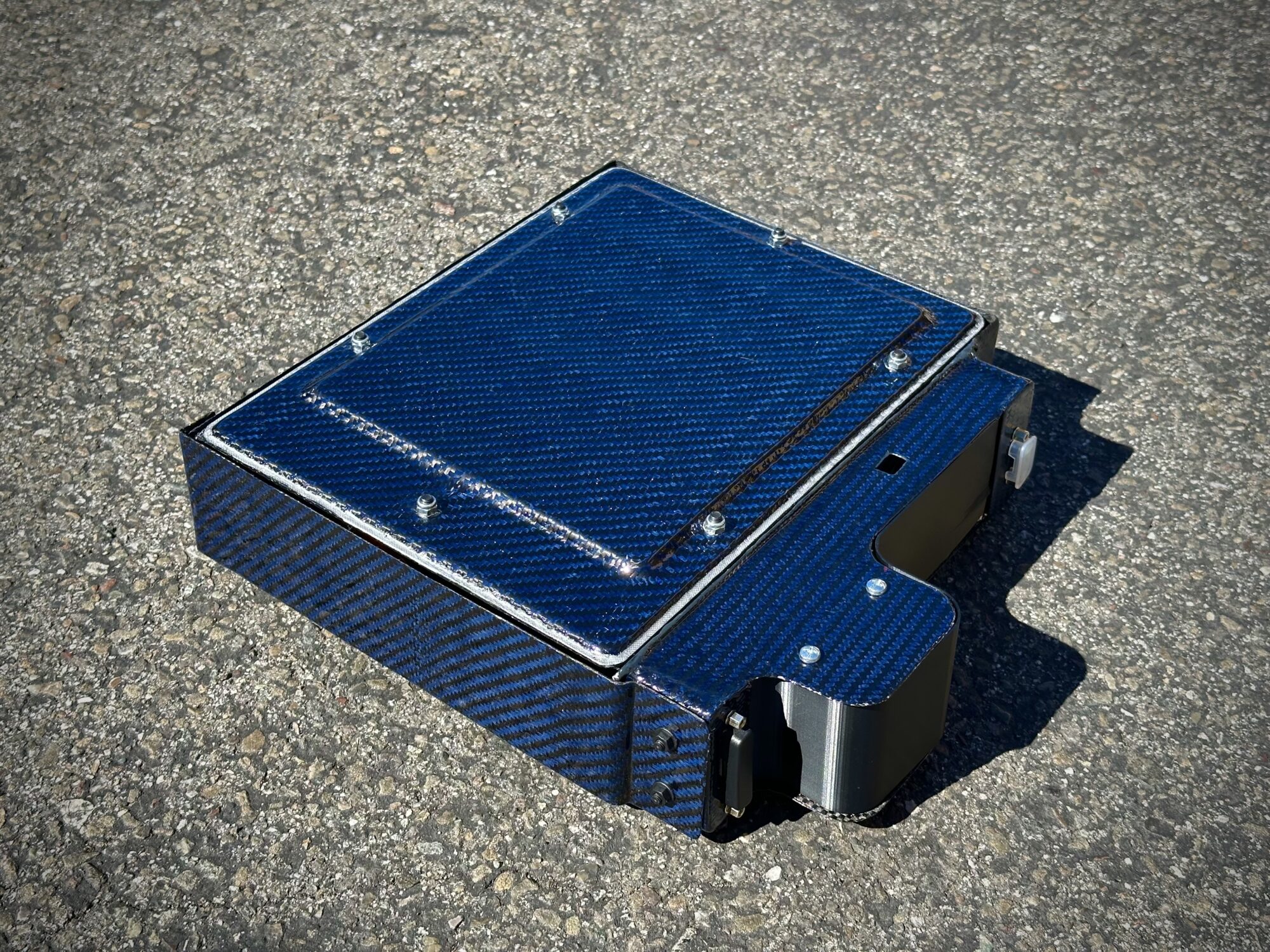 A blue carbon fiber electronic enclosure with screws, resting on outdoor asphalt in bright sunlight, casting shadows.