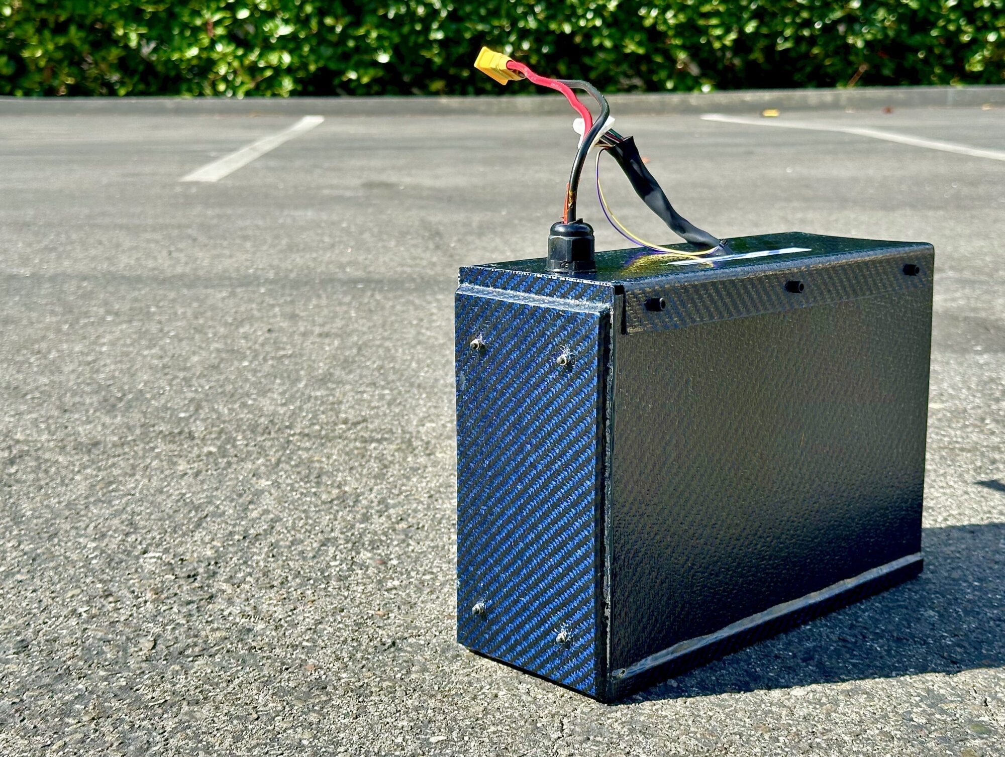 A blue carbon fiber battery pack with wires, placed on asphalt in an outdoor parking lot with greenery behind.