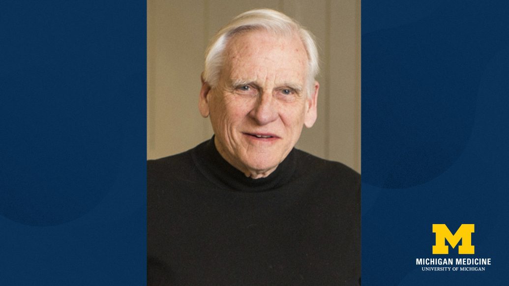 Person with white hair wearing a black sweater, positioned in front of Michigan Medicine and University of Michigan logo.