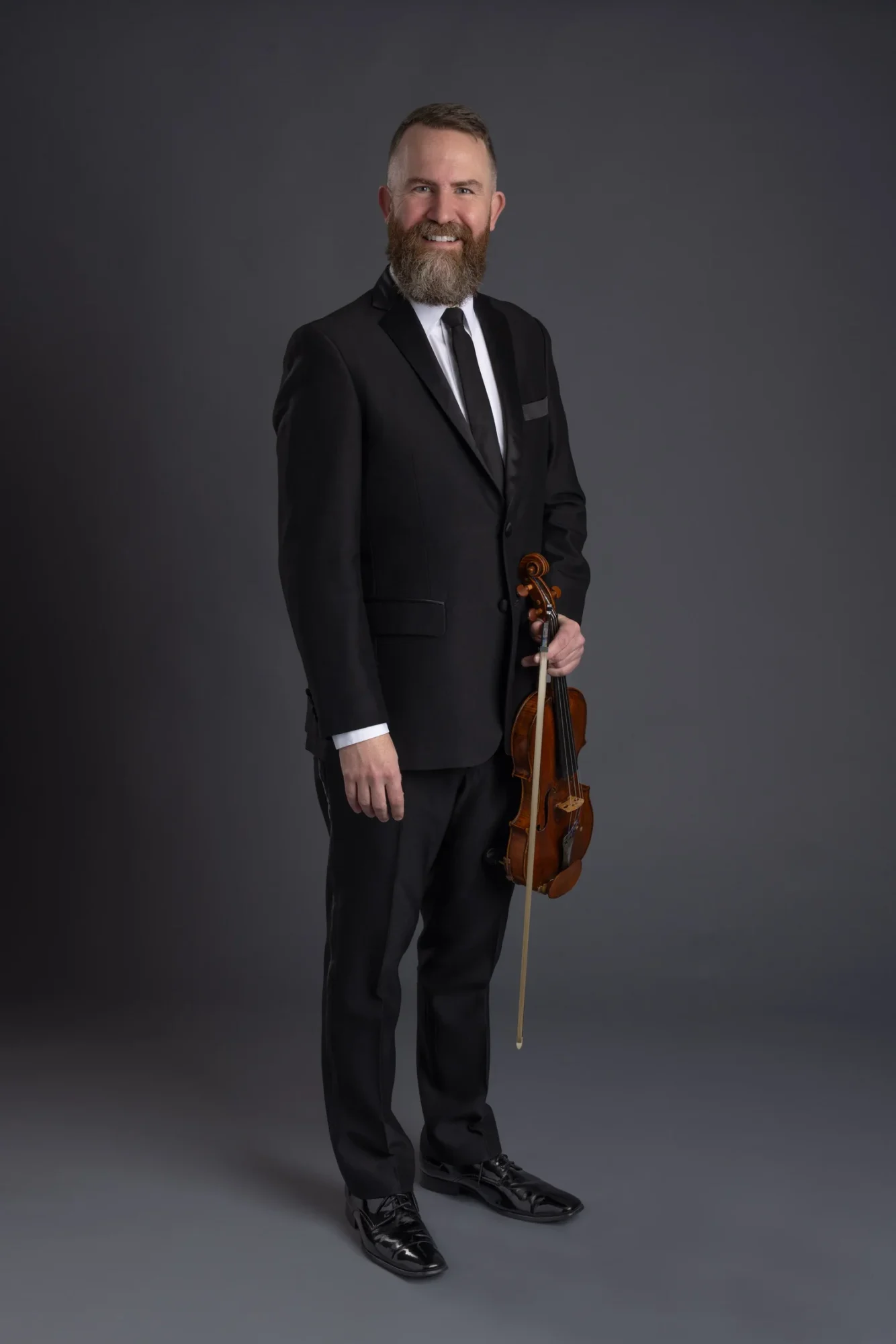 A bearded man standing in formal attire holding a violin and bio in one hand and smiling at the camera.