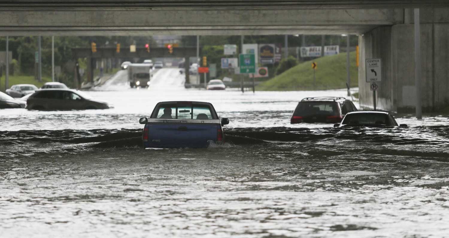 A pickup truck, submerged in water halfway up the driver’s door, is seen from the back attempting to drive beneath an overpass, while nearby cars appear stuck in the flooding.