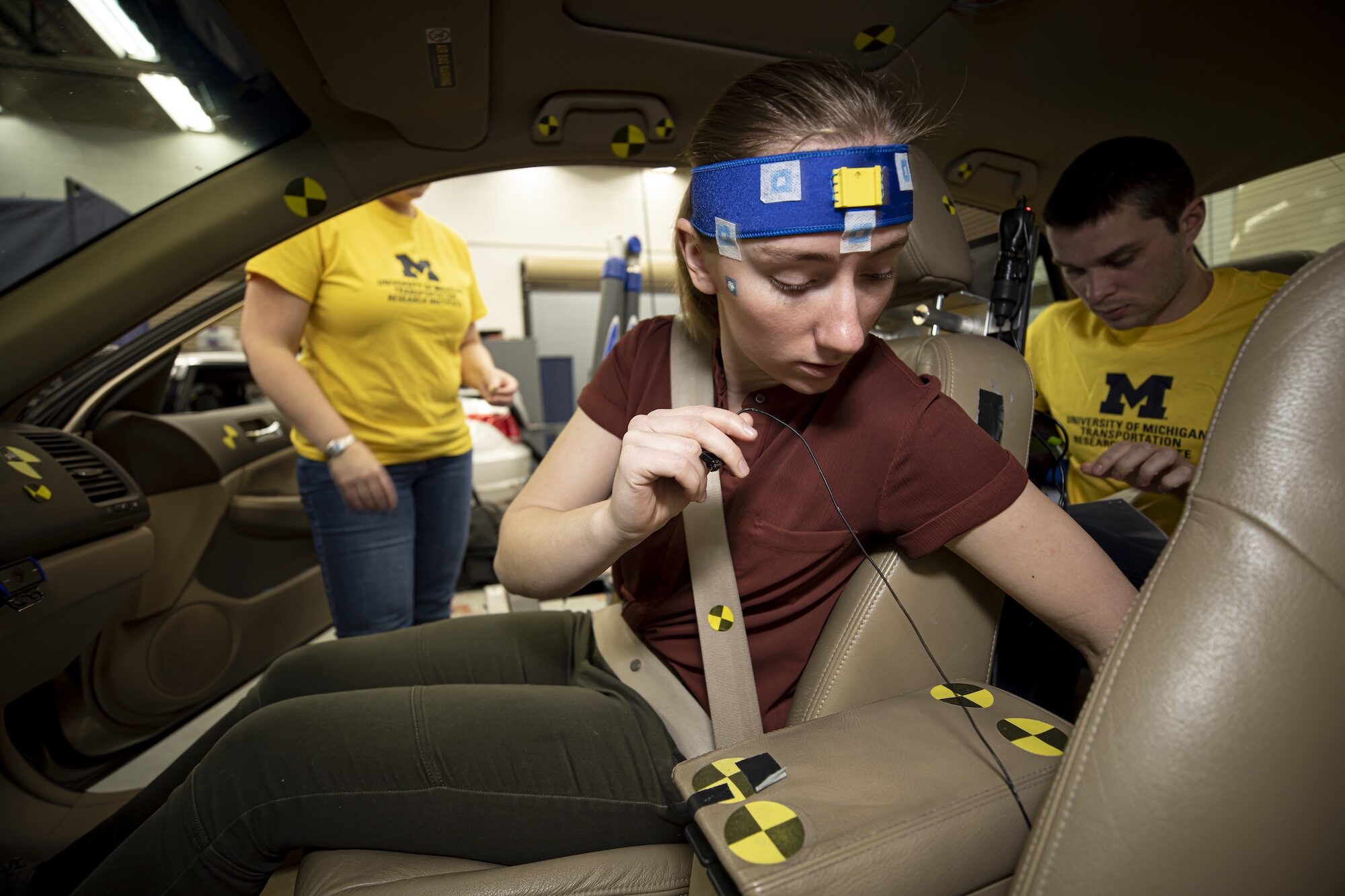 A woman, wearing a sensor strip across her forehead, straps into the passenger seat of a test vehicle while a researcher in the back seat works on a laptop computer.