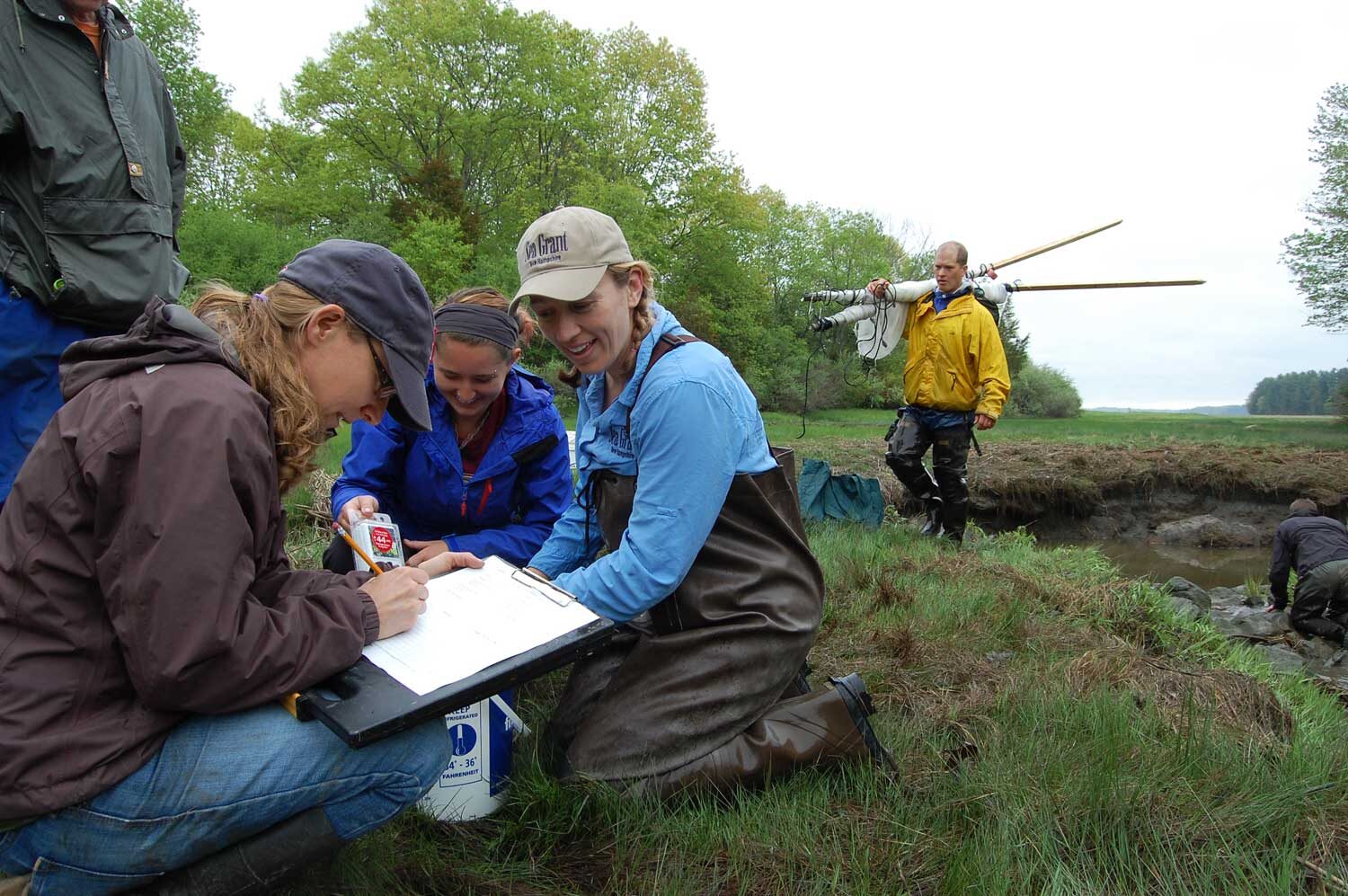 Four NERRS Science Collaborative workers look over documents on a clipboard in a grassy clearing near the water's edge. Another person is carrying gear in the background.