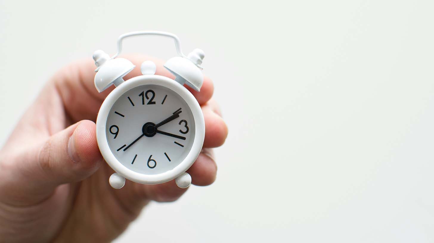 A hand holds a small analog alarm clock in front of a white background.