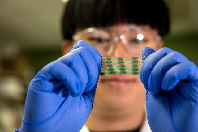 Person wearing blue gloves holds up a transparent square sheet in a laboratory setting, with shelves in background.