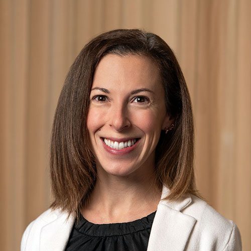 Headshot of a person with straight, shoulder-length brown hair wearing a white blazer over a black top, posed in front of a tan, vertically striped background.