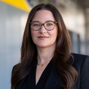 A professional headshot of a person with long wavy brown hair, wearing a black blazer and a necklace, posed indoors against a modern background with a yellow accent in the upper left corner.