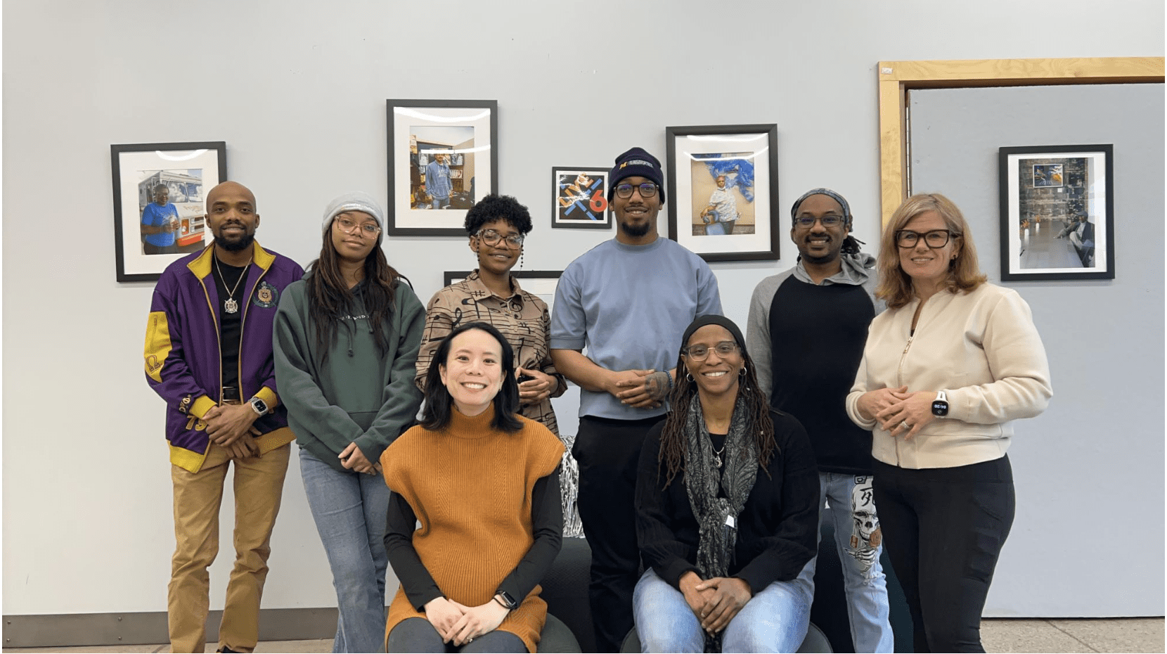 A group of nine people pose together for a photo in front of a wall with framed artwork, with some standing and others seated.