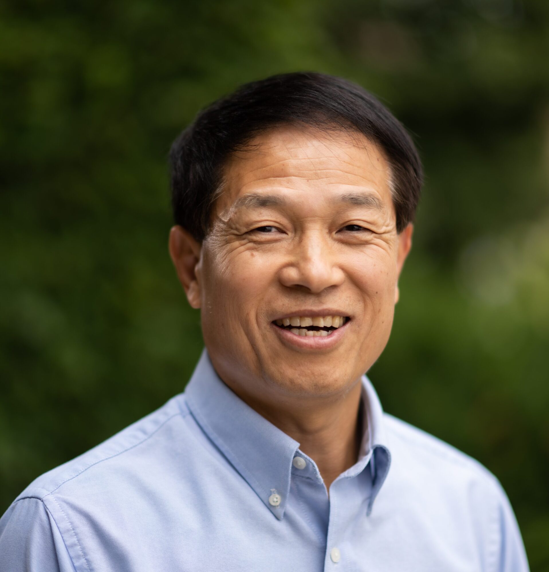 Headshot of a person with short dark hair wearing a light blue button-down shirt, posed outdoors with a green, leafy background.