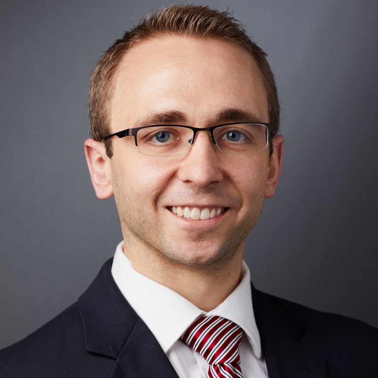 Headshot of a person with short light brown hair, wearing a dark suit jacket, white dress shirt, and a red and white striped tie, set against a plain gray background.