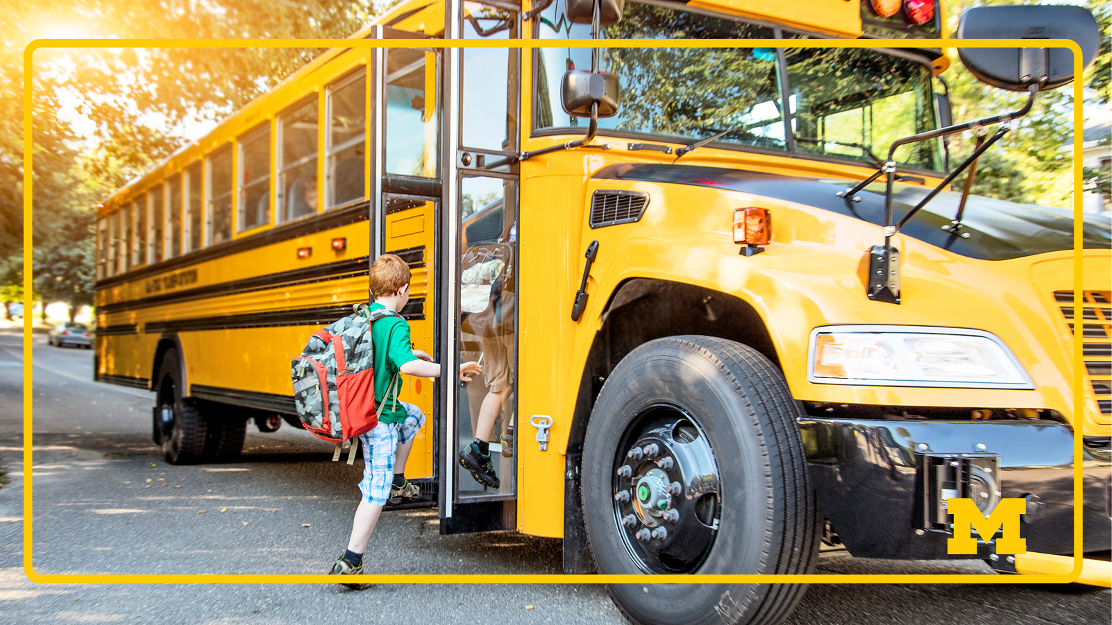 A child wearing a green shirt and a large camouflage backpack with a red pocket is boarding a yellow school bus on a sunny day, with trees and cars lining the residential street in the background.