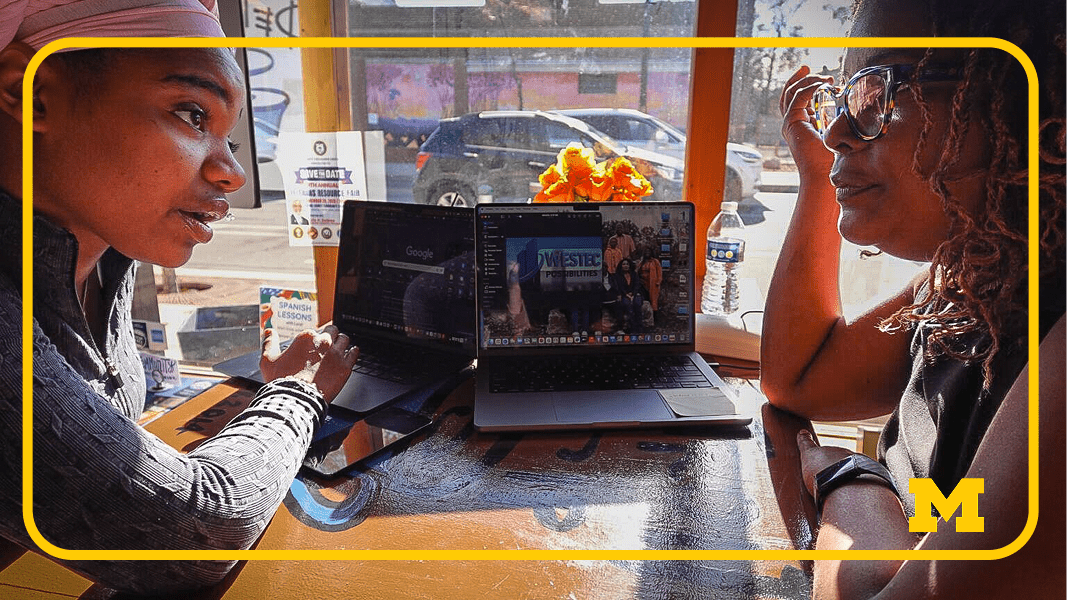 Two people sitting at a table with laptops and water bottles, sunlight streaming through a window with outdoor view.
