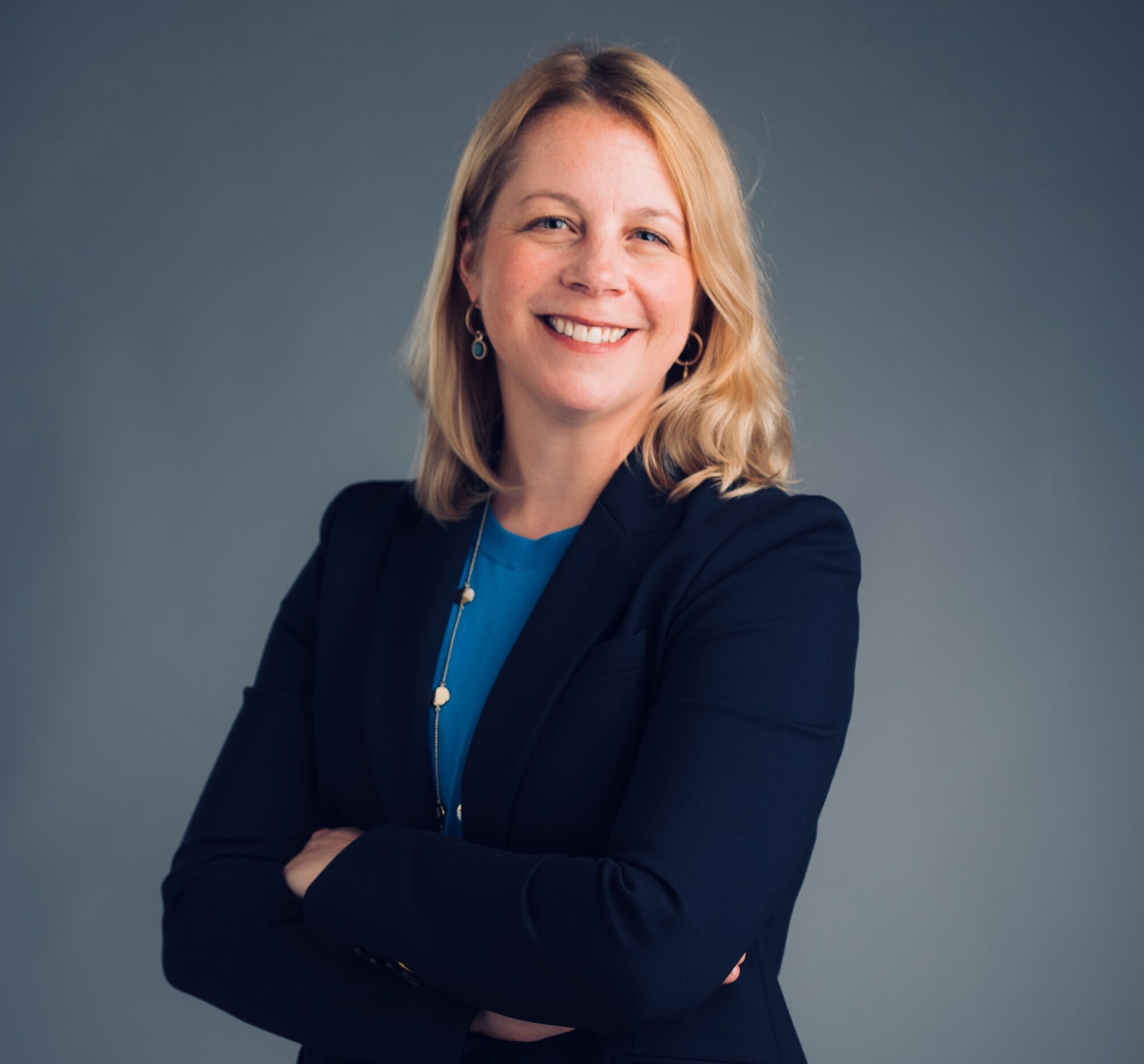 Headshot of a person with shoulder-length blonde hair wearing a dark blazer over a blue top, posing with arms crossed in front of a plain gray background.