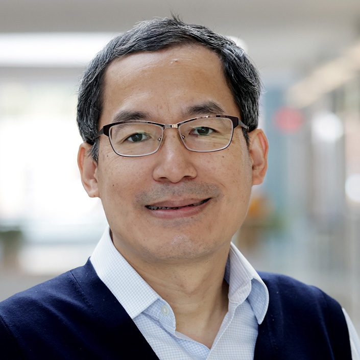 Headshot of a person with short dark hair wearing a light blue collared shirt and a navy blue sweater, standing indoors with a blurred background.