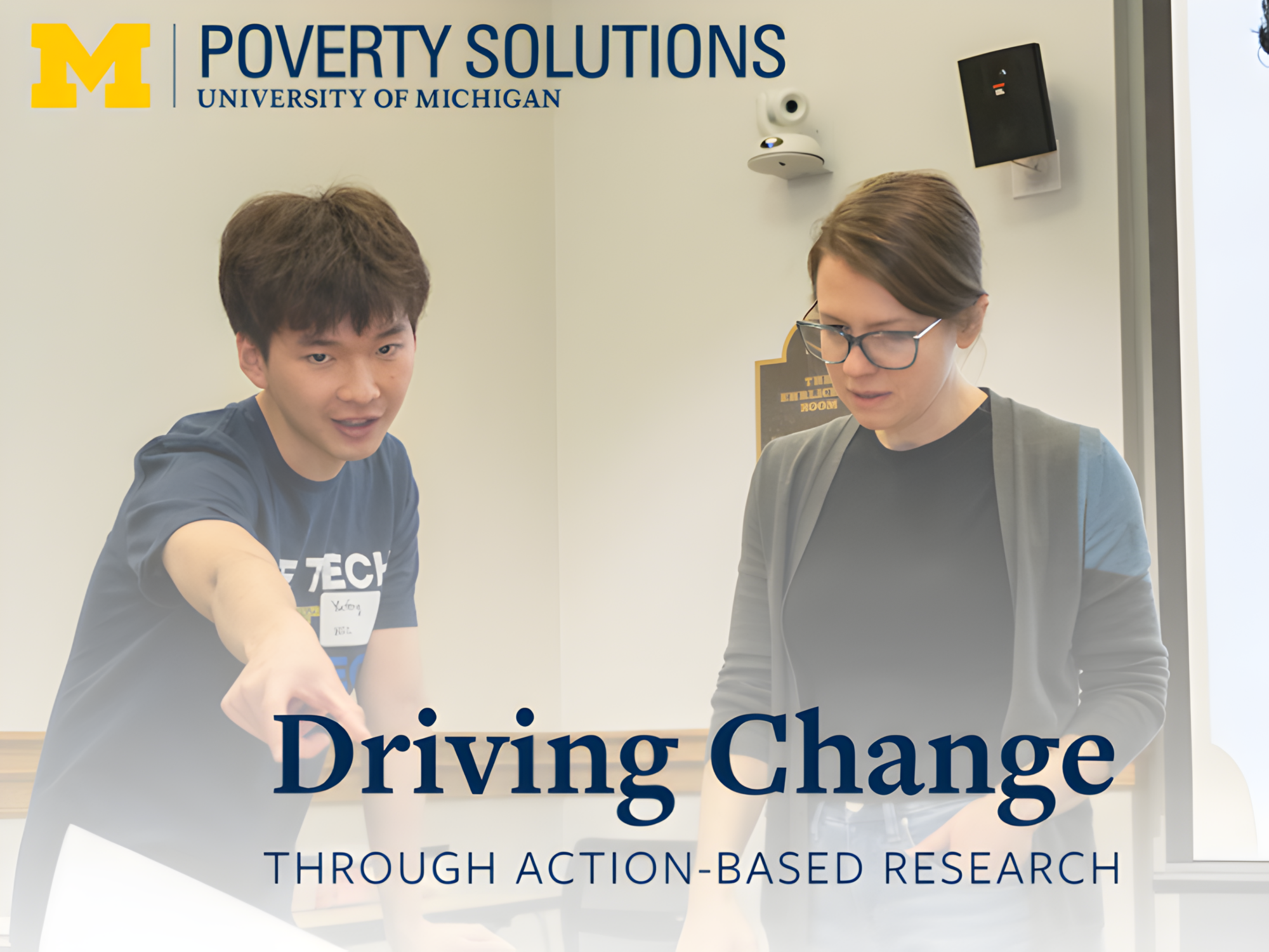 Two people collaborate in a classroom or meeting room with a Poverty Solutions, University of Michigan logo at the top. The text "Driving Change Through Action-Based Research" is prominently displayed across the bottom of the image, emphasizing a focus on community impact and research.