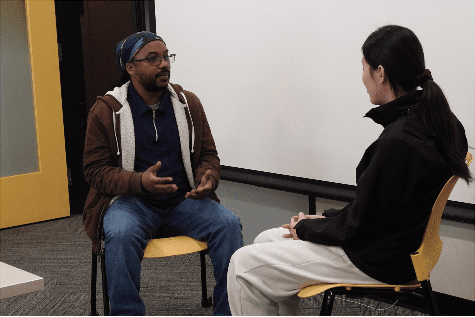 Two people are seated facing each other on yellow chairs in a classroom or meeting room, engaged in conversation. One person gestures with their hands while speaking.