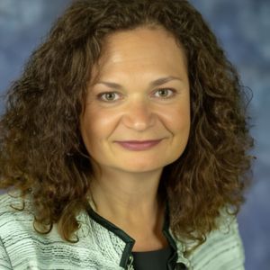 Headshot of a person with curly brown hair wearing a textured light gray jacket with black trim and a long beaded necklace, posed in front of a blue mottled background.