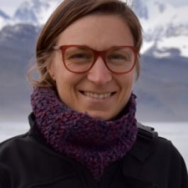 Headshot of a person with light brown hair wearing a dark jacket and a purple knit scarf, standing outdoors with snowy mountains and water in the background.