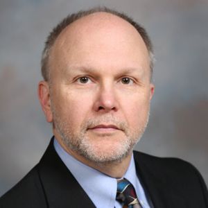Headshot of a person with short gray hair wearing a black suit jacket, light blue dress shirt, and patterned tie, posed in front of a gray mottled background.