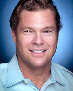 Headshot of a person with light brown, wavy hair wearing a light blue collared shirt, posed against a gradient blue background.